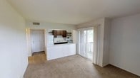 This single-floor residence offers a practical and functional layout centered around an open-concept living and dining area. The spacious main living space is finished with wall-to-wall carpeting and neutral-toned walls, creating a clean and understated ambiance throughout. A half-wall pass-through kitchen connects seamlessly to the living area, providing an open feel while maintaining defined zones. The kitchen features dark wood cabinetry for ample storage, open shelving, a white refrigerator, a gas range, and a sink beneath the pass-through counter, making it both accessible and efficient. Natural light fills the living area through a large sliding glass door dressed with sheer curtains, which opens directly onto a private wooden deck — ideal for outdoor relaxation. The entry door is positioned conveniently off the main living space, offering easy access upon arrival. The overall design emphasizes an open and airy atmosphere, with simple finishes and a neutral palette that provides a versatile backdrop. Ceiling vents indicate a central HVAC system, ensuring year-round comfort throughout the home.