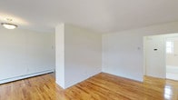 Located at 99 Strathmore Gardens, this single-floor unit offers a clean, functional layout with a bright and consistent aesthetic throughout. Warm-toned hardwood floors run continuously across the living areas and bedrooms, lending a cohesive and inviting feel to the space.

The living area is generously proportioned, featuring crisp white walls, a flush-mount ceiling light, and a large window fitted with blinds that allows ample natural light to fill the room. A wall-mounted air conditioning unit and baseboard heating ensure year-round comfort. The layout includes a partial wall partition that subtly defines separate zones within the open living space.

The kitchen is equipped with white cabinetry accented by light wood trim, laminate countertops, and a full suite of black appliances including a gas range, over-the-range microwave, and refrigerator. The flooring transitions to a durable tile surface, practical for the cooking area.

The bedroom continues the hardwood flooring and includes a closet with bifold doors offering built-in shelving, a window with blinds, and a wall AC unit. The bathroom features full tile surround on the tub-shower combination, chrome grab bars, and a frosted window for natural light and privacy.