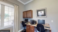 This single-floor business center is thoughtfully designed to provide a functional and comfortable environment for work and leisure. The space is organized into distinct areas that serve different purposes while maintaining a cohesive aesthetic throughout.

The primary workspace features a built-in L-shaped granite-topped desk with rich warm-toned cabinetry below and wall-mounted upper cabinets for additional storage. Multiple computer workstations and a multifunction printer are integrated into the setup, accommodating several users simultaneously. Adjustable ergonomic chairs complete each station. Crown molding and neutral gray walls lend a polished, professional appearance, while plantation shutters and drape-framed windows allow for natural light control.

A separate seating nook offers a more relaxed setting, furnished with dark leather barrel chairs and a glass-topped side table, positioned beside large windows with outdoor views. A tall decorative indoor tree adds a natural element to this corner.

Near the entry, a welcoming station includes a wall-mounted flat-screen television, a single-serve coffee maker, and a dark console table stocked with refreshments. French-paned glass doors connect this area to adjacent spaces, enhancing openness and flow. Carpeted flooring runs throughout, contributing to the warm, inviting atmosphere of the overall space.
