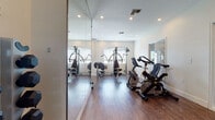 This single-floor fitness facility is thoughtfully designed to support a comprehensive workout experience. The space is organized across multiple interconnected rooms, each dedicated to a specific category of exercise equipment, allowing users to move seamlessly between training areas.

The main exercise area features warm hardwood flooring throughout, crisp white walls, and recessed ceiling lighting that creates a bright, energizing atmosphere. Floor-to-ceiling mirrors are strategically positioned along the walls, visually expanding the space and allowing users to monitor their form during workouts.

The cardio zone is well-stocked with stationary bikes, including upright and recumbent models, as well as treadmills positioned along a wood-paneled accent wall that adds a modern, stylish contrast to the otherwise clean aesthetic. A dedicated strength training section houses a multi-station cable machine by Hoist, ideal for a wide range of resistance exercises, alongside a squat rack with a weight bench for free weight training.

A tiered dumbbell rack stocked with a full range of hex dumbbells provides additional free weight options. Windows fitted with blinds allow natural light to filter in, enhancing the open feel of the space. The layout and variety of equipment make this fitness facility suitable for full-body training routines across all fitness levels.
