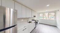 This thoughtfully renovated 3-bedroom, 1-bathroom unit offers a clean, modern aesthetic throughout its single-floor layout. The contemporary kitchen serves as a functional centerpiece, featuring floor-to-ceiling white cabinetry, white quartz countertops, a tile backsplash, and a full suite of stainless steel appliances including a refrigerator, electric range, dishwasher, and range hood. A wide window above the dining area floods the space with natural light and provides views of surrounding greenery. Light gray luxury vinyl plank flooring runs consistently throughout, lending the space a cohesive and polished look. The hallways are bright and open, with recessed lighting and clean white walls that enhance the sense of flow between rooms. Each of the three bedrooms is modestly sized, with the primary bedroom offering a comfortable footprint, and all rooms benefiting from natural light via sliding or picture windows with outdoor views. Select bedrooms include built-in closets with sliding doors for convenient storage. The bathroom is designed with accessibility in mind, featuring grab bars, a vanity with a white countertop, tile flooring, and a window for ventilation. Overall, the space presents a bright, minimalist atmosphere well-suited for efficient, comfortable living.