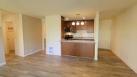 This single-floor space showcases a well-appointed open-concept layout that integrates the kitchen and living area into a cohesive, functional environment. The kitchen is a standout feature, equipped with rich medium-toned wood cabinetry that provides ample storage. Granite countertops with subtle veining add a refined touch, while stainless steel appliances — including a range and built-in microwave — offer modern functionality. A raised peninsula counter serves as both a workspace and a casual dining or entertaining surface, accented by three pendant lights with clean, geometric shades that provide focused task lighting. Recessed ceiling lights further illuminate the kitchen area. The open living space beyond the kitchen benefits from the same warm, neutral wall tones and continuous hardwood-style flooring, which runs throughout and contributes to a cohesive, airy feel. The layout allows for flexible furniture arrangement in the living area. A doorway visible in the background suggests additional rooms, while the overall design balances practicality with an inviting, contemporary aesthetic.