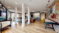 Located at 423 E 36th Street in Charlotte's vibrant Plaza Midwood neighborhood, this single-floor community space is a beautifully designed gathering hub that blends industrial character with refined modern interiors. Wide-plank hardwood flooring runs throughout, anchoring an open-concept layout defined by striking white structural columns that create natural zones without disrupting the spacious flow.

The main lounge area features rich cognac leather seating, layered area rugs, and a large built-in entertainment center with slate-blue cabinetry and open shelving — ideal for relaxed social gatherings. A textured exposed brick accent wall adds warmth and an authentic industrial edge, complemented by a shuffleboard table positioned nearby for recreation. Adjacent to the lounge, a sleek wet bar with white subway tile backsplash and custom cabinetry provides a refined focal point.

A dedicated dining and co-working area is bathed in natural light from floor-to-ceiling windows and sliding glass doors, offering views of the surrounding streetscape. Pendant lighting with brass accents enhances the contemporary aesthetic. An arched doorway leads to additional spaces beyond, adding architectural interest throughout. The overall ambiance is welcoming, sophisticated, and thoughtfully curated for community connection.