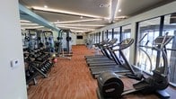 This fitness center is designed to support a comprehensive workout experience within a single, elongated space. The layout is thoughtfully organized, with a row of cardio machines — including treadmills and an elliptical — positioned along a wall of large floor-to-ceiling windows that flood the room with natural light and provide open outdoor views. On the opposite side, a strength training zone features a multi-station cable machine, free weight racks stocked with a full range of dumbbells, and medicine balls for functional training. A wall-mounted flat-screen television is positioned at the far end of the space, visible from multiple workout stations. The ceiling features a distinctive layered design with recessed and accent lighting that adds architectural character while ensuring the room is well-lit at all times. Textured, durable carpeting in warm tones covers the floor throughout, providing both comfort and practicality. Mirrored wall panels near the free weight area enhance the sense of space and allow users to monitor their form during exercise. The overall ambiance is clean, modern, and functional, making it a well-rounded facility suited for a wide variety of fitness routines.