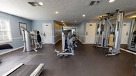 This fitness center offers a thorough workout environment spread across a single floor, divided into distinct zones for both cardiovascular and strength training. The space is finished with durable dark rubber flooring throughout, providing a practical and resilient surface suited to heavy equipment use.

The cardio area features multiple treadmills, an elliptical trainer, and a recumbent stationary bike, arranged alongside a wall-mounted television for entertainment during workouts. Natural light enters through windows fitted with horizontal blinds, and a door provides direct exterior access, contributing to an open, airy feel in this section.

The strength training zone is well-appointed with a variety of Hoist cable and selectorized machines, a multi-function cable station, free weights including dumbbells, and a rowing machine. Large mirrors line portions of the walls, visually expanding the space and allowing users to monitor their form.

Throughout, the walls are painted in a soft blue-gray tone with white chair rail molding, lending a clean, polished aesthetic. Recessed lighting and textured ceilings complete the interior. Additional doors lead to adjacent rooms, suggesting access to restrooms or utility spaces. The overall layout is organized and functional, offering a comprehensive facility suitable for a full range of fitness routines.