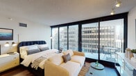 This single-floor urban studio is designed to maximize functionality within an open, well-organized layout. The living and sleeping area is anchored by a queen-sized bed with a tufted navy headboard, positioned to take full advantage of the expansive floor-to-ceiling sliding glass windows that frame striking cityscape views. Natural light floods the interior throughout the day, creating a bright and airy atmosphere. A neutral-toned sofa and a round side table complete the living arrangement, while track lighting overhead provides supplemental illumination in the evenings. The sliding glass doors provide direct access to a private balcony, furnished with seating for outdoor enjoyment.

The kitchen is well-appointed with light maple cabinetry, granite countertops, a dark subway tile backsplash, and a full suite of stainless steel appliances including a gas range, built-in microwave, dishwasher, and refrigerator. Tile flooring throughout the kitchen provides a practical contrast to the dark hardwood floors in the main living area.

The bathroom features a wood-toned vanity with an integrated sink, a large mirror, and neutral wall tones. The overall design of the space balances contemporary urban style with practical, comfortable living.