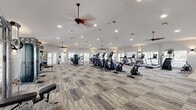 This expansive fitness center is thoughtfully designed to support a wide range of workout routines within a bright, open environment. The space is finished with patterned, textured carpeting in neutral tones accented with blue detailing, lending a modern aesthetic throughout. Recessed lighting and multiple ceiling fans with pendant-style fixtures provide even illumination and comfortable airflow across the entire floor.

Natural light flows generously through numerous windows fitted with white horizontal blinds, as well as multiple glass-paneled entry and exit doors along the perimeter, offering views of the surrounding outdoor areas. The white walls and high ceilings contribute to an airy, open atmosphere.

The cardio zone is well-stocked with a row of treadmills, elliptical trainers, stair climbers, and stationary spin bikes, all arranged to maximize space and ease of movement. A dedicated strength training area features cable machines, multi-function resistance equipment, and a squat rack. Free weights are available alongside a full-length wall mirror, allowing users to monitor their form. Exercise balls and accessories are neatly organized on a rack in a functional stretching and accessory zone. Separate entry points and ample open floor space ensure smooth traffic flow, making this fitness center a complete and practical facility for all fitness levels.