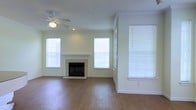 This single-story space centers around a spacious, open living area designed with clean, neutral finishes throughout. The room features warm hardwood-style flooring that extends across the entire space, complementing the crisp white walls and white crown molding that frame the interior. A tile-surround fireplace with a classic white mantel serves as the focal point of the living area, adding a traditional architectural element that anchors the room. Multiple double-hung windows dressed with white horizontal blinds are positioned throughout the space, allowing abundant natural light to fill the interior and create a bright, airy atmosphere. A ceiling fan with an integrated light fixture is centrally mounted, providing both practical ventilation and additional illumination. The overall layout feels open and generously proportioned, with ample space for a variety of furniture arrangements. White baseboards and consistent trim details throughout contribute to a cohesive and polished aesthetic. The neutral color palette and straightforward design create a versatile, move-in-ready environment suitable for a range of uses and personal styles.