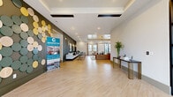 This modern, single-floor community clubhouse offers a generously proportioned and thoughtfully designed gathering space. Light-toned large-format tile flooring flows throughout, unifying the open-concept layout and lending a clean, contemporary aesthetic.

The expansive main lounge serves as the heart of the space, furnished with plush, tufted leather sofas, accent chairs, and decorative throw pillows in a coordinated navy and neutral palette. Bold geometric wall art, a striking dark slatted accent wall, and a gold coffered ceiling detail add layers of visual interest. Recessed lighting and pendant fixtures illuminate the area warmly.

Adjacent to the lounge, the kitchen bar area features a long island with a dark stone countertop, warm wood cabinetry, a marble-style accent wall, a wall-mounted TV, and a dramatic cluster chandelier of globe pendants overhead—an ideal spot for casual socializing or coffee.

Two dining areas provide flexible seating options, each outfitted with solid wood tables and mixed leather and upholstered chairs. One dining room benefits from a striking black-framed glass partition wall that connects to an outdoor courtyard, while large sliding glass doors in the lounge extend the space further outdoors.

The hallway entry features a built-in credenza with curated décor, framed artwork, and black-framed glass double doors, establishing an elegant transition into the broader space.