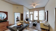 This well-appointed 2-bedroom, 2-bathroom residence is thoughtfully designed to maximize comfort and functionality across a single level. The open-concept living and dining area serves as the heart of the home, featuring warm neutral tones on the walls and ceilings that create a welcoming, airy ambiance. Large windows dressed with sheer curtains flood the living space with abundant natural light, enhancing the sense of openness throughout. A ceiling fan overhead adds both style and practicality to the main living area. The living room is anchored by a plush area rug and complemented by a mix of seating options and a sleek ottoman, making it an ideal space for relaxing or entertaining. Decorative wall art and a tall arched floor lamp add curated, stylish touches. The dining area features a dark reflective table, providing a sophisticated contrast to the lighter palette of the room. A glimpse into one of the bedrooms reveals carpeted flooring and tasteful furnishings, suggesting a comfortable and private retreat. Hardwood-style flooring flows through the main living spaces, tying the design together cohesively. Overall, this residence balances modern aesthetics with livable warmth.