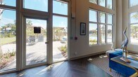 Located at 3320 Riverside Ave in Jacksonville, Florida, this single-story community clubhouse at Rivervue is a well-appointed amenity space designed to serve as a social and recreational hub. The interior immediately impresses with soaring double-height ceilings and an expansive wall of floor-to-ceiling windows and glass doors that flood the space with abundant natural light and frame views of the surrounding palm-lined pool deck and outdoor amenity areas.

The flooring throughout is a striking dark hardwood laid in a classic herringbone pattern, lending the space a sophisticated and cohesive aesthetic. Exposed structural beams and industrial-style track lighting along the ceiling add a contemporary, loft-inspired character to the design.

A central grand staircase with elegant black iron railings serves as a focal architectural feature, flanked by open walkways and leading to an elevated lounge level equipped with a wall-mounted television — ideal for gathering and relaxation. Adjacent to the lounge is a glass-enclosed fitness center, fully equipped with cardio and exercise machines, offering both functionality and visual transparency.

The overall ambiance is bright, airy, and inviting, with carefully placed accent furniture, curated artwork, and decorative lighting that enhance the communal atmosphere. Multiple sets of glass doors provide seamless indoor-outdoor connectivity to the resort-style pool and patio area beyond.