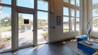 Located at 3320 Riverside Ave in Jacksonville, Florida, this single-story community clubhouse at Rivervue is a well-appointed amenity space designed to serve as a social and recreational hub. The interior immediately impresses with soaring double-height ceilings and an expansive wall of floor-to-ceiling windows and glass doors that flood the space with abundant natural light and frame views of the surrounding palm-lined pool deck and outdoor amenity areas.

The flooring throughout is a striking dark hardwood laid in a classic herringbone pattern, lending the space a sophisticated and cohesive aesthetic. Exposed structural beams and industrial-style track lighting along the ceiling add a contemporary, loft-inspired character to the design.

A central grand staircase with elegant black iron railings serves as a focal architectural feature, flanked by open walkways and leading to an elevated lounge level equipped with a wall-mounted television — ideal for gathering and relaxation. Adjacent to the lounge is a glass-enclosed fitness center, fully equipped with cardio and exercise machines, offering both functionality and visual transparency.

The overall ambiance is bright, airy, and inviting, with carefully placed accent furniture, curated artwork, and decorative lighting that enhance the communal atmosphere. Multiple sets of glass doors provide seamless indoor-outdoor connectivity to the resort-style pool and patio area beyond.