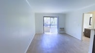 This single-floor space is characterized by its clean, minimalist layout and an abundance of natural light. The main living area features smooth hardwood flooring that reflects sunlight throughout the room, enhancing the sense of openness and airiness. Crisp white walls and white baseboards contribute to a bright, neutral aesthetic that provides a versatile backdrop for any furnishing style.

A prominent sliding glass door serves as a central architectural feature, fitted with vertical blinds that allow for adjustable light control. The door opens directly onto a balcony, offering views of mature, lush greenery outside — a notable detail that brings a natural element into the living space. The balcony access also provides a seamless indoor-outdoor connection.

A wall-mounted air conditioning unit is positioned near the sliding door, offering a practical climate control solution integrated into the wall. The room's layout is straightforward and spacious, with no obstructions, making it highly adaptable for various furniture arrangements. Overall, the space presents a light-filled, functional environment with clean finishes and pleasant outdoor connectivity.