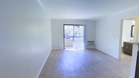 This single-floor space is characterized by its clean, minimalist layout and an abundance of natural light. The main living area features smooth hardwood flooring that reflects sunlight throughout the room, enhancing the sense of openness and airiness. Crisp white walls and white baseboards contribute to a bright, neutral aesthetic that provides a versatile backdrop for any furnishing style.

A prominent sliding glass door serves as a central architectural feature, fitted with vertical blinds that allow for adjustable light control. The door opens directly onto a balcony, offering views of mature, lush greenery outside — a notable detail that brings a natural element into the living space. The balcony access also provides a seamless indoor-outdoor connection.

A wall-mounted air conditioning unit is positioned near the sliding door, offering a practical climate control solution integrated into the wall. The room's layout is straightforward and spacious, with no obstructions, making it highly adaptable for various furniture arrangements. Overall, the space presents a light-filled, functional environment with clean finishes and pleasant outdoor connectivity.