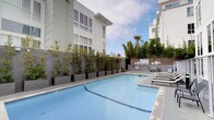 This outdoor amenity space is designed to offer residents a resort-style experience within an urban residential setting. The centerpiece is a generously sized swimming pool with clear turquoise water, surrounded by a smooth concrete pool deck that provides ample space for lounging and recreation. Chaise lounge chairs are positioned poolside, offering comfortable spots to relax and enjoy the open sky above.

Adjacent to the main pool sits a dedicated spa, or hot tub, with tiled surrounds and a stainless steel handrail for easy entry and exit. Seating is available nearby, making the spa area a comfortable retreat. Safety signage and equipment, including a life ring, are prominently displayed throughout.

The pool area is enclosed by sleek glass panel railings that maintain an open, airy feel while providing a clear boundary. Lush potted trees and greenery line the perimeter, adding a natural touch and a sense of privacy from the surrounding multi-story buildings. A palm tree and additional mature plantings further enhance the resort-like atmosphere.

The space benefits from abundant natural light and open sky views, creating a bright and welcoming outdoor environment. The overall design is clean, modern, and well-maintained, reflecting a high standard of residential amenity living.