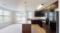 Located at 15 Abbey Ln in Danbury, Connecticut, this single-floor residence offers a clean, modern layout designed for comfortable everyday living. The interior is finished with a neutral color palette of crisp whites and warm cream tones throughout, creating a bright and welcoming atmosphere.

The kitchen serves as a central highlight, featuring rich dark-espresso cabinetry, light countertops, black appliances including a gas range and dishwasher, and a central island with pendant lighting overhead. Wood-look flooring runs through the kitchen area, while large windows allow natural light to fill the adjoining living space, which transitions to plush carpeting.

The living area is open and generously proportioned, offering a seamless connection to the kitchen and providing ample room for a variety of furniture arrangements. A wide hallway connects the main living areas to the private quarters, with a built-in HVAC vent and access panel visible along the corridor.

The bedroom is spacious and carpeted, accompanied by a dedicated walk-in closet with built-in shelving and hanging rods. The full bathroom is well-appointed with warm tile flooring, a dark-toned vanity with a wide countertop and mirror, and a tiled tub-shower combination with chrome fixtures. The overall design is functional and cohesive, reflecting a thoughtfully planned single-level floor plan.