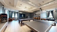 Located at 12 Digital Way in Maynard, MA, this thoughtfully designed game and entertainment lounge delivers a compelling mix of recreational amenities within a single, well-appointed floor. The space showcases a sophisticated interior design that blends industrial and contemporary elements, featuring tray ceilings with recessed and track lighting, light hardwood-style flooring, and a combination of textured tile and bold plaid-patterned wallcoverings that create distinct zones within the open layout.

At the heart of the room, a full-size billiards table and a ping-pong table take center stage beneath a stylish chandelier, while a dark stone accent wall with a mounted flat-screen television and cue rack provides a focal point for the main gaming area. A bar-height seating area with warm leather stools offers a relaxed spot to socialize. Adjacent to the main area, a lounge nook features plush seating, a shuffleboard table, and curated artwork displayed in a backlit gallery wall, adding a refined touch to the space.

A standout feature is the dedicated indoor golf simulator room, complete with floor-to-ceiling impact screens, turf flooring in multiple tones, acoustic wall panels, and a leather lounge chair — ideal for immersive play and relaxation. Large windows throughout invite natural light, enhancing the vibrant yet welcoming ambiance of this exceptional recreational space.