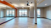 Located at 100 District Dr in Asheville, NC, this single-floor fitness studio offers a well-appointed and functional space designed for a variety of exercise activities. The main studio area is expansive and open, featuring light hardwood-style flooring that runs throughout the entire space, contributing to a clean and modern aesthetic. Full-length mirrored walls line one side of the studio, accented by a bold orange accent wall that adds energy and visual interest to the room. Large floor-to-ceiling windows wrap around the space, flooding the interior with natural light and offering views of the surrounding Asheville landscape, including glimpses of the Blue Ridge Mountains in the distance. Ceiling fans and linear fluorescent light fixtures ensure the space remains comfortable and well-lit during use. Structural columns are integrated thoughtfully throughout the open floor plan without impeding usability. A dedicated equipment zone houses a tiered dumbbell rack, stability balls, and additional fitness accessories, keeping the main floor clear for group classes or individual workouts. An interior glass-paneled door connects to an adjacent room, allowing for flexible use of the overall space. Informational displays and signage are mounted near the entrance, providing users with schedules and facility details. The overall ambiance is bright, motivating, and professionally designed for active use.
