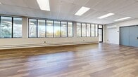 Located at 283 Pharmacy Ave in Toronto's east end, this single-floor commercial space offers a generously sized, open-concept layout well-suited for a variety of uses, from event hosting and fitness studios to office or community programming. The interior is defined by wide-plank hardwood flooring that runs throughout, lending warmth and continuity to the expansive floor plan. A prominent wall of large, black-framed windows stretches along one side of the space, flooding the interior with natural light and providing scenic views of the surrounding treetops. The drop ceiling is fitted with a grid of recessed fluorescent panel lights and decorative diffuser vents, ensuring consistent, even illumination across the entire area. White-painted walls provide a clean, neutral backdrop, while baseboard heating units run beneath the window wall for year-round comfort. Multiple doorways with steel-framed doors offer access points throughout the space, including clearly marked emergency exits. The open floor plan affords significant flexibility in furniture arrangement and room configuration. A small seating area with sofas and a dining set with chairs is visible, illustrating the space's adaptability. The overall ambiance is bright, clean, and functional, making this a highly versatile commercial space in a convenient Toronto location.