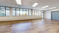 Located at 283 Pharmacy Ave in Toronto's east end, this single-floor commercial space offers a generously sized, open-concept layout well-suited for a variety of uses, from event hosting and fitness studios to office or community programming. The interior is defined by wide-plank hardwood flooring that runs throughout, lending warmth and continuity to the expansive floor plan. A prominent wall of large, black-framed windows stretches along one side of the space, flooding the interior with natural light and providing scenic views of the surrounding treetops. The drop ceiling is fitted with a grid of recessed fluorescent panel lights and decorative diffuser vents, ensuring consistent, even illumination across the entire area. White-painted walls provide a clean, neutral backdrop, while baseboard heating units run beneath the window wall for year-round comfort. Multiple doorways with steel-framed doors offer access points throughout the space, including clearly marked emergency exits. The open floor plan affords significant flexibility in furniture arrangement and room configuration. A small seating area with sofas and a dining set with chairs is visible, illustrating the space's adaptability. The overall ambiance is bright, clean, and functional, making this a highly versatile commercial space in a convenient Toronto location.