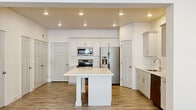 This two-story home offers a thoughtfully designed layout that balances functional living spaces with a bright, contemporary aesthetic. The kitchen serves as the heart of the main floor, showcasing shaker-style cabinetry in a soft gray-white finish, a large center island with a crisp white quartz countertop, and a full suite of stainless steel appliances including a French door refrigerator with ice and water dispenser, a range with microwave above, and a dishwasher. Wood-look tile flooring runs throughout the kitchen and main living area, adding warmth and durability. Recessed lighting illuminates the space efficiently throughout.

A carpeted living area adjoins the kitchen in an open-concept arrangement, with a ceiling fan overhead providing comfort. A staircase with plush carpet leads to the upper level, where multiple bedrooms offer generous proportions, natural light through large windows with blinds, and soft neutral carpeting. Each bedroom is well-lit, with ceiling fixtures and clean, neutral walls.

The bathrooms feature quartz countertops, undermount sinks, shaker-style cabinetry with brushed nickel hardware, and large mirrors. A dedicated laundry room on the upper level houses a full-sized top-load washer and dryer alongside open shelving for storage. The home's clean lines and neutral palette create a fresh, move-in-ready atmosphere throughout.