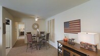 This single-story space presents a warm and inviting dining area anchored by a round, dark-toned bistro-style table surrounded by matching wrought iron chairs with rich wooden accents. Soft, neutral-colored carpeting runs throughout, complementing the clean white walls and creating a cohesive, understated aesthetic. A ceiling fan with a built-in light fixture overhead provides both comfort and ambient illumination. Decorative touches lend the space a distinct rustic, Americana character — a weathered wooden American flag artwork commands attention on one wall, while a starburst sunburst mirror adds dimension nearby. Linen-toned wall hangings provide additional texture against the neutral backdrop. A wooden console or media unit along the wall offers practical storage while displaying decorative accents including a glass vase and vintage-style scale. A hallway visible in the background hints at additional rooms beyond, suggesting a well-connected and functional layout. Overall, the space carries a comfortable, lived-in charm with a cohesive rustic-patriotic design theme throughout.