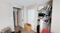 This single-floor unit offers a practical and functional layout across its living areas. The bedroom is generously sized, featuring a large window with vertical blinds that allows natural light to fill the room, a textured popcorn ceiling, and a sliding mirrored closet that adds depth to the space. The bedroom also includes a dedicated area suitable for a home office setup. Built-in open shelving provides additional storage alongside a separate closet accessible through a standard interior door. The hallway connects the bedroom to other areas of the unit and features multiple doors, offering access to additional storage or utility spaces. The kitchen is compact yet well-equipped, with warm wood-toned upper and lower cabinetry, granite-look countertops, a stainless steel refrigerator, and a gas range. A sink positioned beneath the cabinetry completes the kitchen's practical layout. The flooring transitions from carpet in the bedroom and hallway areas to hardwood-style flooring in the kitchen, adding a distinction between the living and cooking zones. Neutral warm beige wall tones carry throughout the unit, creating a cohesive and understated aesthetic across all areas.