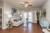This single-floor residence is designed with a cohesive coastal-inspired color palette of soft neutrals, whites, and teal accents that carry throughout each space. The living area is bright and welcoming, featuring wood-style flooring and a set of French doors that provide natural light and direct access to an outdoor area, creating a seamless indoor-outdoor connection.

The kitchen is efficiently laid out with rich, dark espresso cabinetry contrasted by light stone-style countertops and a classic tile backsplash. Stainless steel appliances, including a refrigerator, range, and microwave, are integrated throughout, and a built-in fold-down desk area provides a convenient workspace within the kitchen footprint.

The bedrooms are carpeted and comfortably scaled, each featuring a ceiling fan and windows dressed with light curtains that allow natural light to filter in softly. The primary bedroom connects to a spacious walk-in closet outfitted with wire shelving along multiple walls and washer/dryer hookups, adding functional storage and laundry convenience.

The bathroom includes wood-style flooring, dark cabinetry with a light countertop vanity, a single sink, and a separate tiled soaking tub and shower combination, offering a clean and practical layout throughout.