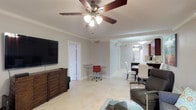 This single-floor residence offers a thoughtfully designed open-concept layout that seamlessly connects the living, dining, and kitchen areas into one cohesive and functional space. The living area is anchored by a wooden media console with a wall-mounted flat-screen television, complemented by comfortable seating and a glass coffee table. A five-blade ceiling fan with an integrated light fixture provides both comfort and illumination while adding a classic touch to the room. Crown molding runs consistently throughout, lending a polished and refined aesthetic to the interiors. Light-toned tile flooring flows across the entire space, creating a clean and bright foundation that ties each area together. A compact workspace with a glass desk and ergonomic chair is thoughtfully positioned within the living area, offering a dedicated spot for productivity without disrupting the overall flow. The dining area features a sleek dark table paired with white high-back chairs, providing a stylish contrast to the light surroundings. The kitchen, visible in the background, is equipped with dark cabinetry, stainless steel appliances, and pendant lighting, combining functionality with contemporary design. The overall ambiance is bright, airy, and well-organized.