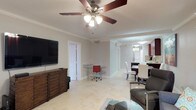 This single-floor residence offers a thoughtfully designed open-concept layout that seamlessly connects the living, dining, and kitchen areas into one cohesive and functional space. The living area is anchored by a wooden media console with a wall-mounted flat-screen television, complemented by comfortable seating and a glass coffee table. A five-blade ceiling fan with an integrated light fixture provides both comfort and illumination while adding a classic touch to the room. Crown molding runs consistently throughout, lending a polished and refined aesthetic to the interiors. Light-toned tile flooring flows across the entire space, creating a clean and bright foundation that ties each area together. A compact workspace with a glass desk and ergonomic chair is thoughtfully positioned within the living area, offering a dedicated spot for productivity without disrupting the overall flow. The dining area features a sleek dark table paired with white high-back chairs, providing a stylish contrast to the light surroundings. The kitchen, visible in the background, is equipped with dark cabinetry, stainless steel appliances, and pendant lighting, combining functionality with contemporary design. The overall ambiance is bright, airy, and well-organized.