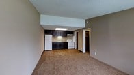 This single-floor space offers an efficient and practical open-concept layout that seamlessly connects the living area and kitchen. The living space features wall-to-wall carpeting in a warm beige tone, complemented by neutral taupe-colored walls that create a cozy, understated ambiance throughout.

The kitchen is positioned along the back wall and is outfitted with dark espresso-toned cabinetry that provides a striking contrast against the lighter surroundings. It includes a white refrigerator, a white electric range, and a built-in sink with a countertop that features subtle stone-like detailing. An overhead range hood is installed above the stove, and a recessed skylight panel in the kitchen area provides additional brightness to the workspace.

The textured popcorn ceiling spans the entire space, adding a classic touch to the interior. A doorway visible from the main living area leads to what appears to be a walk-in closet or storage area with warm interior lighting, suggesting practical and well-considered storage solutions. The layout is designed for straightforward functionality, making efficient use of the available space. Overall, the unit presents a clean, move-in-ready environment with a simple and cohesive design aesthetic.