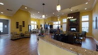 This single-floor community space is designed to be both functional and inviting, with an open-concept layout that seamlessly connects its various areas. At the forefront is a curved reception counter finished in warm granite, serving as the central hub of the space. Bold yellow walls give the interior an energetic and cheerful character, complemented by rich hardwood-style flooring that runs throughout.

The space boasts soaring ceilings with crown molding details, and an abundance of natural light streams in through multiple large windows positioned along the perimeter. Recessed lighting and stylish pendant fixtures provide additional illumination throughout the day and evening.

To one side, a relaxed lounge area features comfortable dark upholstered seating arranged around a striking floor-to-ceiling stone fireplace with a rustic wood mantel, creating a warm and cozy gathering spot. On the opposite side, a dining and activity area is furnished with tables and chairs, ideal for meetings or social gatherings. A large flat-screen television and additional seating make this a versatile multipurpose area.

A decorative sculptural figure near the reception counter adds a playful, welcoming touch to the overall ambiance. The thoughtful combination of warm tones, natural materials, and open sightlines gives this space an approachable and community-oriented feel.