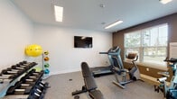 Located at 7479–7485 Outer Loop in Louisville, KY, this single-floor fitness center is a well-equipped exercise facility designed to support a range of workout routines. The space features durable speckled rubber flooring throughout, providing both comfort and functionality for various types of physical activity.

The cardio area is outfitted with a treadmill, a recumbent stationary bike, and a spin/cycling bike, offering users multiple options for cardiovascular training. A weight bench is also present, complementing the dedicated free weights section, which includes a multi-tiered dumbbell rack stocked with a full set of hex dumbbells. A medicine ball rack holds a colorful assortment of weighted balls alongside stability balls, catering to functional and core training needs.

Multiple wall-mounted flat-screen televisions are positioned throughout the space, allowing users to stay entertained during workouts. The facility is illuminated by recessed and flush-mounted fluorescent light panels in the ceiling, while a window along one wall admits natural light and views of the surrounding outdoor greenery. A glass-panel entry door provides direct exterior access and reinforces the bright, open feel of the space.

The interior walls are finished in a two-tone scheme—light gray on the upper portions and a deeper charcoal brown accent on the lower sections—giving the fitness center a clean, modern appearance. Overall, the space is thoughtfully organized to maximize usability within its footprint.