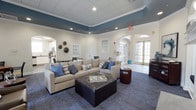 Located at 8101 Fonthill Court in Langhorne, PA, this single-floor space offers a thoughtfully designed interior that blends comfort with refined style. The main lounge area serves as the centerpiece of the space, featuring a plush neutral-toned sectional sofa accented with blue and striped throw pillows, complemented by a dark leather ottoman that doubles as a coffee table. The room is anchored by a rich blue tray ceiling with recessed lighting, creating a striking contrast against the crisp white crown molding and walls. Decorative wall plates add an artistic touch to the otherwise clean aesthetic. Arched doorways and passages lend an architectural elegance throughout, while French-style glass doors invite natural light and provide views to an outdoor area. The flooring features a patterned blue-toned carpet that ties the color palette together cohesively. Adjacent to the lounge, a well-appointed kitchen is visible, offering white cabinetry and modern appliances, ideal for gatherings and events. The overall ambiance is welcoming and polished, making this a versatile and inviting communal environment suited for relaxation and social interaction alike.
