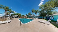 Located at 902 SH-499 Loop in Harlingen, Texas, this single-story outdoor facility is centered around a generously sized swimming pool set within a well-maintained, landscaped grounds. The pool area features a broad concrete sun deck surrounding the water, furnished with multiple white lounge chairs positioned to capture the abundant South Texas sunshine. The facility is framed by tall, mature palm trees and established shade trees that lend a tropical character to the setting.

Distinctive teal and turquoise-painted single-story buildings border the pool area, providing a cohesive and vibrant aesthetic throughout the property. A covered pavilion structure on one side of the pool offers a shaded gathering space, while additional seating areas are distributed across the deck for flexible outdoor use. The grounds are enclosed by a perimeter fence, ensuring privacy and security for users of the space.

Visible pool equipment is neatly situated along the building exterior, and signage indicates proper hygiene protocols are in place. Patches of green lawn surround portions of the pool deck, adding a natural contrast to the hardscape. The overall ambiance is relaxed and resort-like, benefiting from the warm Harlingen climate and open sky views, making this an inviting outdoor recreational destination suited for community or hospitality use.