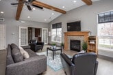 This single-floor community space is thoughtfully designed to accommodate a variety of gatherings and activities. The main lounge area is an architectural highlight, featuring soaring vaulted ceilings accented by warm, exposed wood beams and ceiling fans. Light hardwood flooring runs throughout, complementing the neutral gray walls and recessed lighting. A stone-surround fireplace anchors one end of the room beneath a wall-mounted television, while large windows allow ample natural light to fill the space. Comfortable seating arrangements, including a sectional sofa and leather accent chairs, create an inviting atmosphere. French doors connect to additional areas, including a dedicated media room with a vaulted ceiling, large projection screen, and plush seating on carpeted floors—ideal for screenings or private events. A well-appointed kitchen offers generous honey-toned cabinetry, dark granite countertops, stainless steel appliances including a side-by-side refrigerator and dishwasher, and tile flooring. A transition corridor with a cozy seating nook and a "gather" wall sign provides a welcoming entry point. An additional flex room with rich hardwood flooring and oversized windows rounds out the layout, offering space for meetings or smaller gatherings.