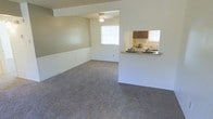 This single-story space features an open living area finished with wall-to-wall carpet in a neutral tone, contributing to a quiet and comfortable atmosphere. The living room is generously sized, with a ceiling fan providing both lighting and ventilation. Natural light enters through a window fitted with horizontal blinds, brightening the otherwise neutral-toned walls. A distinctive architectural element is the pass-through window that connects the living area to the kitchen, enhancing the sense of openness between the two spaces while maintaining a degree of separation. The kitchen features dark wood cabinetry and a countertop that extends slightly to serve as a bar-style ledge, practical for casual dining or serving. The kitchen also benefits from its own window, ensuring adequate natural light. The overall design is straightforward and functional, with clean lines and a neutral color palette throughout. The layout promotes an efficient flow between the living and kitchen areas, making the space well-suited for everyday living.