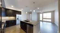 This single-floor space showcases a thoughtfully designed open-concept layout that seamlessly connects the kitchen and living areas. The kitchen is a standout feature, centered around a spacious island with a granite countertop, an undermount double sink, and a built-in dishwasher. Sleek dark cabinetry provides generous storage and creates a striking contrast against the light walls and wood-tone flooring that runs throughout the space. Stainless steel appliances, including a French door refrigerator with an ice and water dispenser, add a polished, modern touch. Pendant lighting hangs above the island, providing focused task lighting while contributing to the overall contemporary aesthetic. Recessed lighting throughout ensures the space is well-illuminated. The living area flows naturally from the kitchen and is bathed in natural light from large windows and a glass door that opens to an outdoor area, offering pleasant exterior views. A ceiling fan overhead enhances comfort in this gathering space. A hallway visible from the main area leads to additional rooms, completing this functional and well-appointed single-level layout.