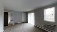 This single-floor space presents a straightforward and functional layout centered around a open living area. The room features wall-to-wall carpeting in a neutral beige tone, contributing to a quiet and understated atmosphere. Gray-painted walls run throughout, lending the space a clean, modern feel.

A large sliding glass door serves as a prominent architectural feature, fitted with vertical blinds that allow for adjustable natural light. The door provides direct access to the outdoors and brightens the interior considerably. Baseboard heating units are positioned along multiple walls, ensuring efficient climate control throughout the space.

The ceiling features a classic drop-tile panel design, maintaining a clean overhead aesthetic. A solid white interior door is visible along one wall, leading to an adjacent area of the unit. A light switch and door hardware are the primary wall fixtures noted in this section of the space.

Overall, the space offers a practical and neutral environment, well-suited for customization. The combination of natural light from the sliding door, carpeted flooring, and perimeter heating creates a livable and functional interior ready for occupancy.