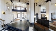 This impressive lobby space is defined by its soaring double-height ceilings and towering white columns that anchor the room with a sense of grandeur. An open mezzanine level with dark-railed balconies wraps above, adding vertical drama and architectural depth. The interplay of crisp white walls and bold dark trim creates a striking contrast that emphasizes the space's historic character.

The flooring features a classic black-and-white tile pattern that reinforces the formal elegance of the entrance. Ornate Moorish-style pendant lanterns hang at varying heights, casting warm, ambient light throughout and serving as distinctive decorative focal points. Large multi-pane windows near the entrance allow natural light to filter in, brightening the expansive interior.

A heavily carved dark wood settee with richly striped upholstery provides seating near an intricately detailed dark wood reception or front desk structure, both showcasing exceptional craftsmanship. A brass floor lamp adds a refined, boutique-like warmth to the seating area. The overall ambiance is one of timeless, classical elegance with an institutional or hospitality-oriented character, blending historic architectural detail with functional public space design.