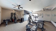 This single-floor fitness center is designed to support a comprehensive exercise routine in a practical, functional setting. The space is divided into two connected zones, each serving a distinct workout purpose. The cardio area is outfitted with treadmills, an elliptical trainer, and a recumbent bike, positioned near a window that allows natural light to brighten the room. A wall-mounted television provides entertainment during workouts. The second zone houses a multi-station cable weight machine alongside a stability ball, catering to strength training needs. A dark accent wall adds a modern visual element, contrasting with the neutral beige tones throughout. The carpeted flooring provides a degree of cushioning underfoot, while multiple ceiling fans with integrated lighting ensure consistent air circulation and adequate illumination. A wall-mounted air conditioning unit supports climate control. Framed wall art and minimal décor contribute a clean, organized aesthetic. The textured ceiling and straightforward layout reflect a utilitarian design focused on functionality, making this fitness center a convenient and accessible amenity for regular use.