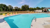 This outdoor swimming pool area offers a well-appointed recreational space designed for community use. The generously sized pool features clearly marked depth indicators, including a designated no-diving shallow end, and is equipped with stainless steel entry handrails for safe and easy access. The water is maintained to a clear, bright turquoise condition, reflecting the open sky above.

The surrounding concrete pool deck provides ample space for lounging, furnished with multiple yellow and orange chaise lounge chairs positioned along the perimeter. A red market umbrella with accompanying seating offers a shaded area for those seeking relief from direct sunlight. A black wrought iron fence encloses the entire pool area, providing security and defined boundaries. Mature trees visible beyond the fence contribute a natural backdrop to the space.

An adjacent utility building serves as the operational hub for the pool facility. Inside, shelved storage rooms are stocked with a comprehensive inventory of pool chemicals, cleaning supplies, and maintenance materials. A separate mechanical room houses the pool's filtration and plumbing infrastructure, including a sand filter and an interconnected network of PVC piping and valves essential for water circulation and treatment.