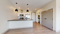 Located at 2885 Detroit Ave in Cleveland's vibrant Ohio City neighborhood, this single-floor unit offers a thoughtfully designed open-concept layout with clean, contemporary finishes. The kitchen serves as a centerpiece of the space, featuring a large island with a dark granite countertop, undermount sink, and seating potential. White shaker-style cabinetry pairs with a subway tile backsplash, while stainless steel appliances — including a French door refrigerator, range, and microwave — complete the modern aesthetic. Statement black pendant lights hang above the island, adding a stylish contrast to the bright, neutral palette. Light-toned luxury vinyl plank flooring flows seamlessly throughout the entire unit, unifying the open kitchen and living areas. The living room is generously sized and bathed in natural light through multiple large windows that frame views of the Cleveland cityscape. A central structural column adds an architectural element to the room. The bathroom features large-format tile surrounding a soaking tub and shower combination, complemented by warm wood-toned flooring and gold accent hardware. Ample closet storage is provided by full-height double doors. The overall ambiance is bright, airy, and move-in ready.