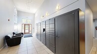 This single-floor amenity space is thoughtfully divided into two distinct functional areas designed to serve the needs of a residential community. The first area is a welcoming lobby and package room, characterized by soaring ceilings, large clerestory windows, and warm wall sconces that fill the space with abundant natural light. Light-toned tile flooring runs throughout, and a wall of modern dark metal package lockers provides a convenient, secure solution for parcel delivery. A pair of leather armchairs near the entrance creates a comfortable seating nook, while double-door entry with a welcome mat provides an inviting threshold to the outdoors. The second area is a dedicated business center, accessible through a connecting doorway. This room features warm wood-tone luxury vinyl plank flooring, recessed linear lighting, and generous desk workspace along two walls, accommodating multiple computer workstations and monitors. Ergonomic task chairs line the desks, making it ideal for productivity. A leather sofa and loveseat offer additional seating, lending a relaxed atmosphere to the work environment. Together, the two areas create a cohesive, practical, and comfortable amenity suite that balances utility with an inviting aesthetic.