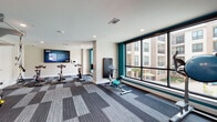 This single-floor fitness center is thoughtfully designed to accommodate a comprehensive range of workout styles, organized across several distinct training zones that flow seamlessly into one another. The space features a contemporary aesthetic, with patterned modular carpet tiles in shades of grey, blue, and brown laid in varying geometric configurations throughout, providing visual definition between areas.

The cardio zone is positioned along a wall of large, floor-to-ceiling windows that frame views of an adjacent courtyard and surrounding building, flooding the area with natural light. This section houses elliptical machines, stationary bikes, and a rowing machine, complemented by a wall-mounted television for entertainment. Teal accent walls add a bold, energetic color contrast throughout the space.

The strength training area is expansive and well-stocked, featuring a multi-station cable and functional training rig, a punching bag, weight benches, and a full dumbbell rack. Floor-to-ceiling mirrors line the walls, visually amplifying the space and supporting proper form during exercise.

A dedicated functional training zone includes TRX suspension trainers, stability balls, and a wall-mounted storage rack. Glass-panel partitions separate select zones while maintaining an open sightline. Recessed lighting provides consistent, even illumination throughout. The overall design is modern, clean, and purposeful.