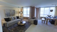 This well-appointed single-floor space showcases a thoughtfully designed living area that balances comfort and contemporary style. The living room is a standout feature, anchored by large windows dressed with elegant patterned curtains that allow generous amounts of natural light to fill the room while offering views of the surrounding exterior. The space is finished with warm, neutral tones — rich brown accent walls contrast tastefully against light-colored walls and ceilings adorned with crown molding. Plush wall-to-wall carpeting in a soft cream tone runs throughout, adding warmth underfoot. The living area is furnished with a spacious grey sofa accented by decorative pillows, complemented by a pair of neutral-toned armchairs. A distinctive metallic oval coffee table serves as a focal point, resting atop a dark floral-patterned area rug that adds texture and depth to the room. Accent pieces including a tall floor lamp, a round black side table, and abstract wall art contribute to the curated, sophisticated aesthetic. The open layout promotes a sense of airiness and flow, making the space feel expansive and welcoming. Overall, the design strikes a balance between modern elegance and everyday livability.