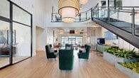 Located at 726 Dixon Ct in Exton, PA, this expansive single-floor facility is thoughtfully designed to serve as a modern community and amenity hub. The centerpiece is a dramatic double-height lobby showcasing soaring ceilings, oversized rattan and drum pendant lighting, and light oak hardwood flooring that flows throughout. A curved mezzanine with sleek black steel railings overlooks the main gathering area, accessible via a bold open-riser staircase, adding vertical architectural interest to the space.

The main lounge features clusters of deep emerald velvet armchairs arranged to encourage conversation, while vertical wood slat partitions define zones without sacrificing openness. A glass-walled private office provides visual transparency and separation, and a built-in display shelving unit with teal backing serves as a striking design focal point in one of the seating areas.

A dedicated lounge room offers large floor-to-ceiling windows with outdoor views, a wall-mounted television, leather bench seating, and direct access to an exterior patio. The fully equipped fitness center includes strength and cardio equipment, floor-to-ceiling mirrors, and large windows that invite natural light. An elevator provides additional accessibility throughout the facility.