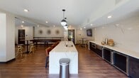 Located at 214 Seventh Street West in Saint Paul, Minnesota, this contemporary community amenity space is designed to offer residents a well-appointed gathering environment with distinct zones for relaxation, entertainment, and socializing. The space is unified by warm hardwood-style flooring and a neutral color palette that gives the interior a modern, cohesive feel.

A standout feature is the large central kitchen island topped with striking white marble-veined quartz countertops, illuminated by a trio of globe pendant lights. The kitchen area is equipped with built-in appliances, a beverage cooler, and ample bar seating with leather stools, making it ideal for catered events or casual get-togethers. Dark cabinetry along the back wall provides a bold contrast to the light stone backsplash, while a wall-mounted television adds entertainment functionality.

Adjacent to the kitchen is a relaxed lounge area anchored by a dramatic floor-to-ceiling cartographic mural depicting the Saint Paul cityscape — a nod to the building's local identity. Comfortable seating, including a neutral tufted sofa and a bold yellow accent chair, invites residents to unwind. A shuffleboard table positioned toward the back of the space adds a recreational dimension, complemented by high-top bar tables and stools for more informal social interaction. Recessed lighting throughout ensures a bright, welcoming ambiance.