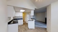 This single-floor residence is designed with an open-concept layout that seamlessly connects the kitchen and living area, creating a spacious and functional living environment. The kitchen is well-appointed with crisp white cabinetry, dark countertops, and a stainless steel sink. A peninsula with a cooktop serves as both a practical cooking space and a natural divider between the kitchen and the adjacent living area. A full-size refrigerator and additional upper cabinetry provide ample storage throughout. The kitchen features wood-look flooring that adds warmth and durability underfoot, transitioning smoothly into plush carpeting in the living area. One of the standout architectural features is the vaulted ceiling, which rises dramatically and is highlighted by a skylight that draws in natural light, enhancing the open and airy ambiance of the space. A hallway visible from the kitchen leads to additional areas of the home. The living area benefits from the generous ceiling height, contributing to an expansive feel throughout. The overall design palette, featuring neutral gray tones, white finishes, and natural textures, creates a clean, contemporary aesthetic that is both inviting and versatile.