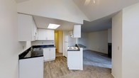 This single-floor residence is designed with an open-concept layout that seamlessly connects the kitchen and living area, creating a spacious and functional living environment. The kitchen is well-appointed with crisp white cabinetry, dark countertops, and a stainless steel sink. A peninsula with a cooktop serves as both a practical cooking space and a natural divider between the kitchen and the adjacent living area. A full-size refrigerator and additional upper cabinetry provide ample storage throughout. The kitchen features wood-look flooring that adds warmth and durability underfoot, transitioning smoothly into plush carpeting in the living area. One of the standout architectural features is the vaulted ceiling, which rises dramatically and is highlighted by a skylight that draws in natural light, enhancing the open and airy ambiance of the space. A hallway visible from the kitchen leads to additional areas of the home. The living area benefits from the generous ceiling height, contributing to an expansive feel throughout. The overall design palette, featuring neutral gray tones, white finishes, and natural textures, creates a clean, contemporary aesthetic that is both inviting and versatile.